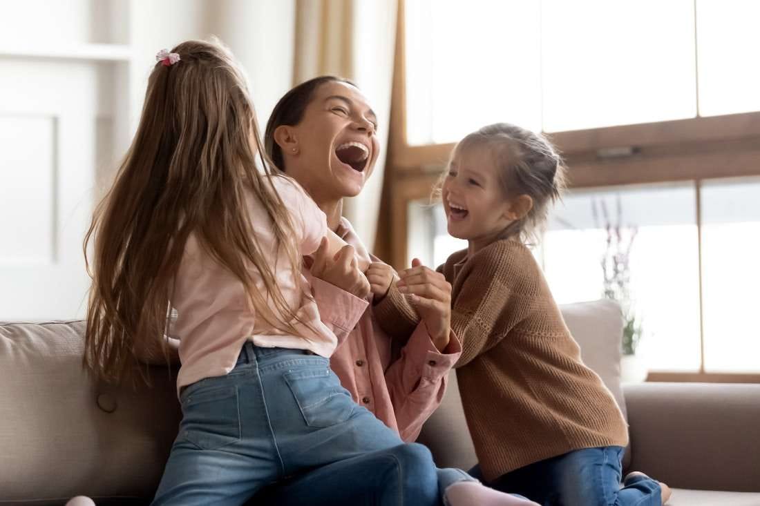 A mother laughing with her daughters in a bright living room.